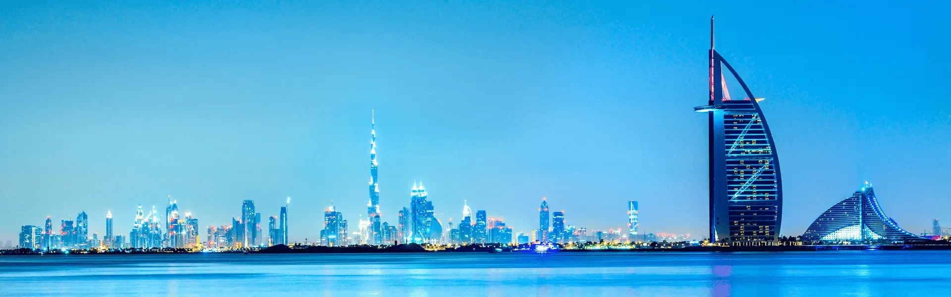 Dubai skyline at night with the sail-shaped Burj Al Arab and illuminated skyscrapers reflected on calm water.