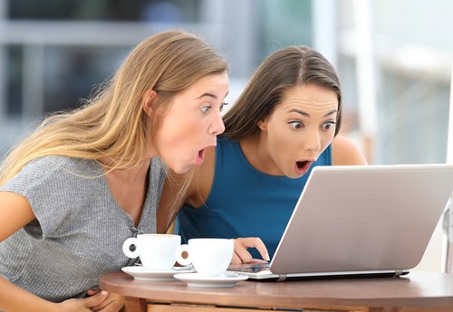 Two women looking amazed to a laptop while having coffee outside