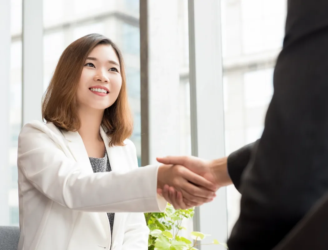 A professional businesswoman in a white blazer smiles warmly while shaking hands with a colleague in a bright modern office, marking a successful meeting or new partnership.