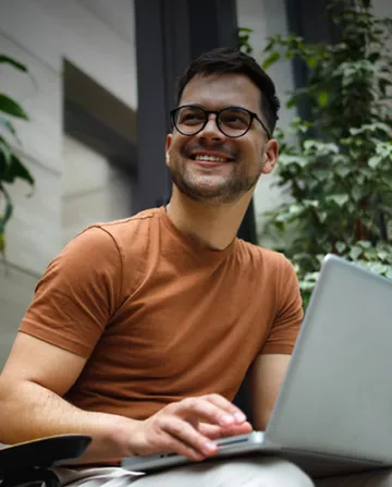 Smiling man wearing glasses and a brown t-shirt, sitting outdoors with greenery in the background, typing on a laptop.