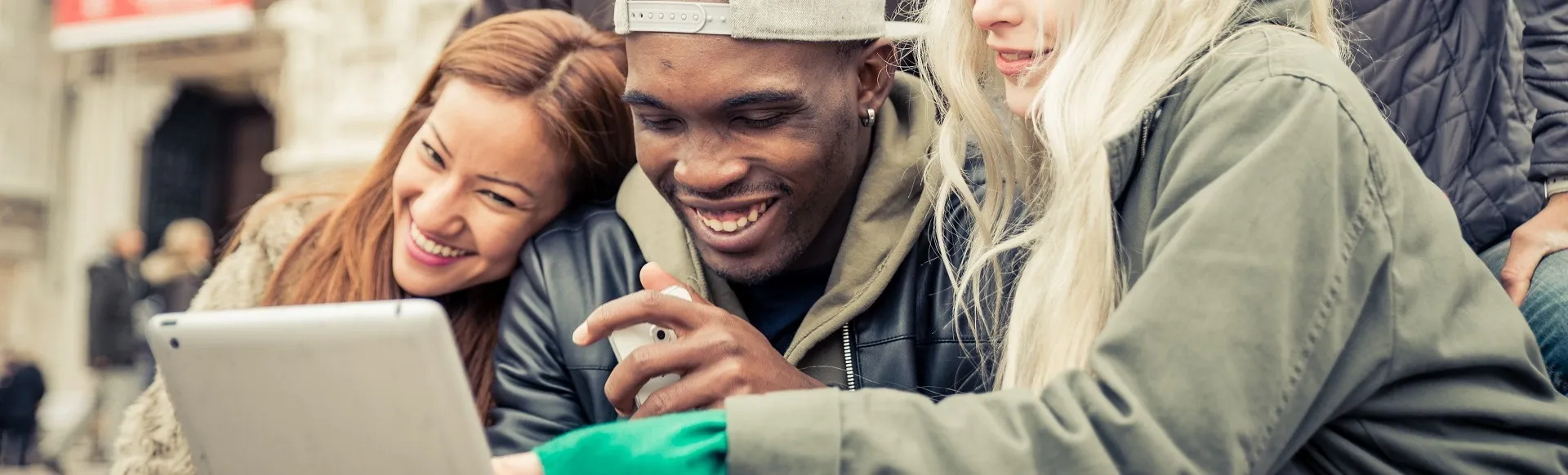 Friends smiling while looking at a tablet together outdoors.