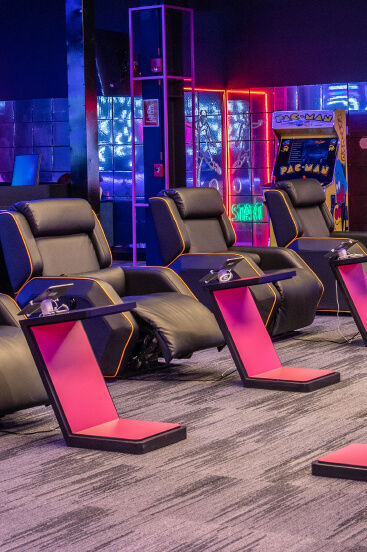 Sala de juegos con sillones reclinables, escritorios rosas y máquinas arcade iluminadas por luces de neón al fondo.