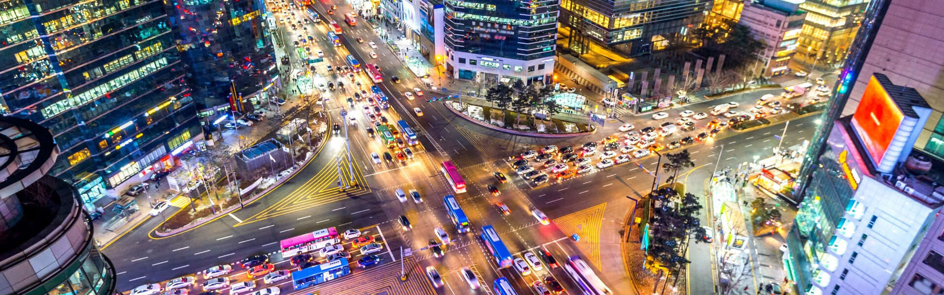 분주한 도시 교차로의 항공 뷰로, 수많은 자동차와 버스가 도로를 오가고 있습니다. 주변의 불빛이 켜진 고층 건물들이 활기찬 밤의 도시 풍경을 더욱 돋보이게 합니다.
