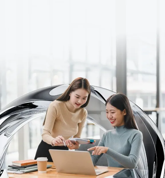Two professional women collaborate happily at work, discussing ideas on a laptop in a bright modern office.