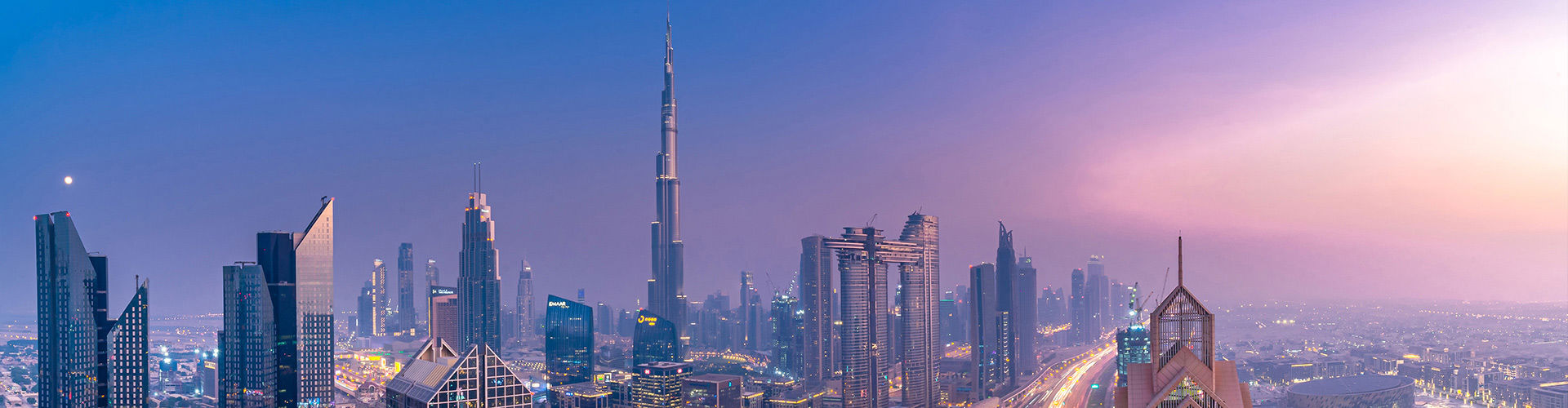 Panoramic view of Dubai cityscape at dusk featuring Burj Khalifa and surrounding modern skyscrapers with illuminated buildings and roads.