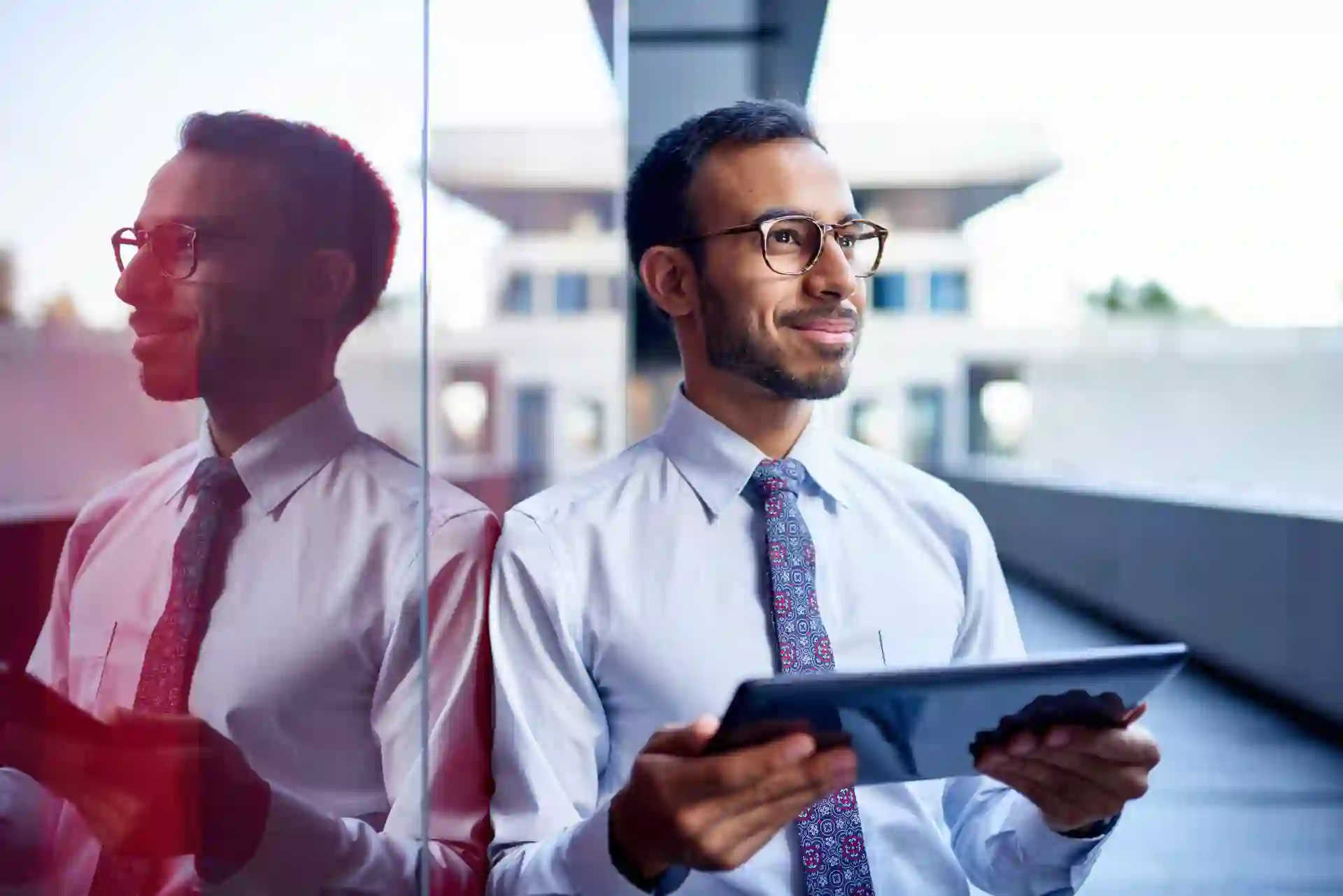Businessman holding tablet while leaning against a glass wall reflecting his image