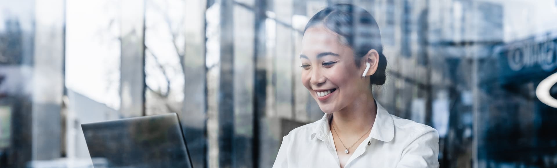 Smiling Asian professional working on her laptop inside a modern glass-walled coworking space.