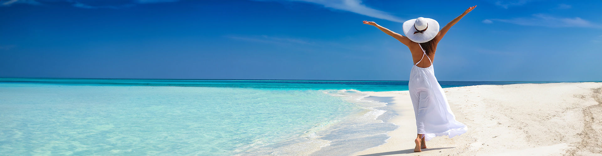 Woman in a white summer dress and wide-brimmed hat enjoying a tropical beach with turquoise water and clear blue sky.