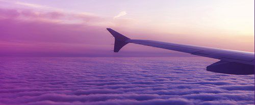 a wing extends against a vibrant sunset sky, flying above a blanket of clouds in shades of purple and pink, creating a serene, colorful aerial view.