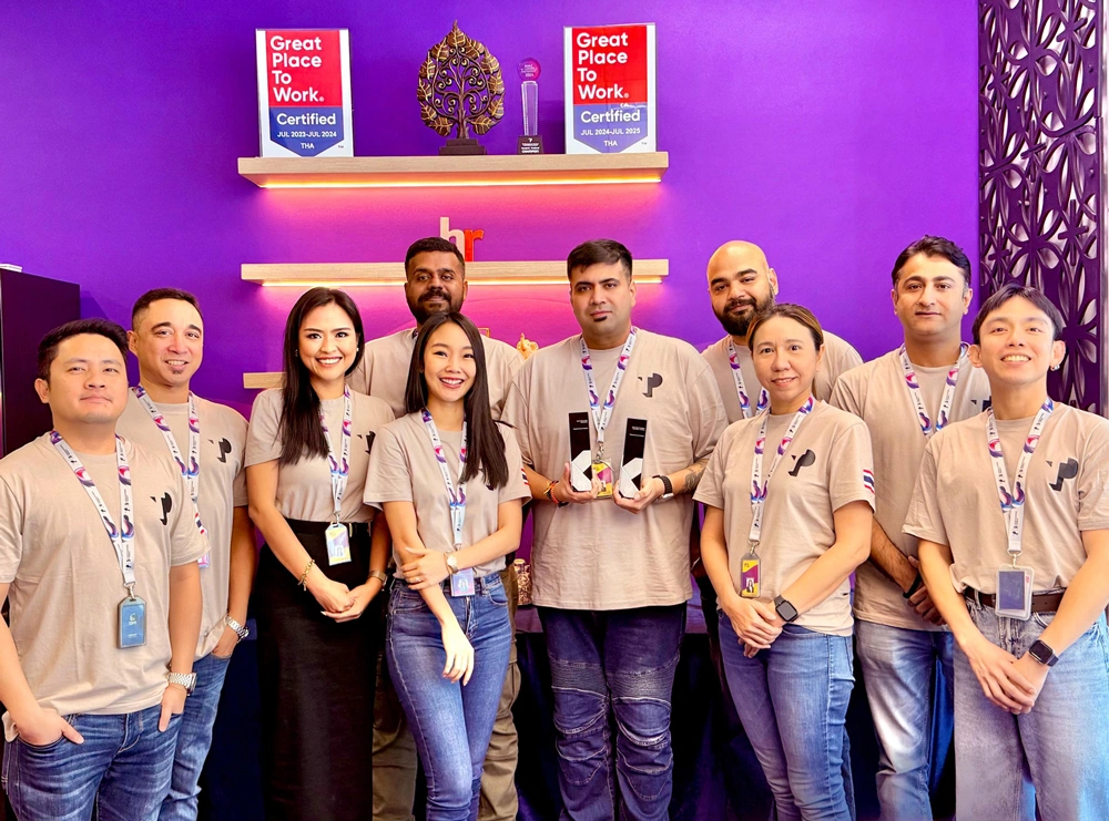 A diverse team in matching branded shirts poses proudly, celebrating a milestone with awards at a corporate event.