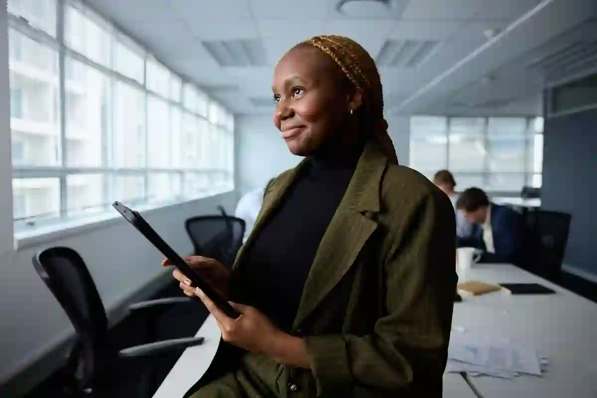 Happy African employee holding a tablet and looking up while sitting at a meeting desk inside a bright meeting room