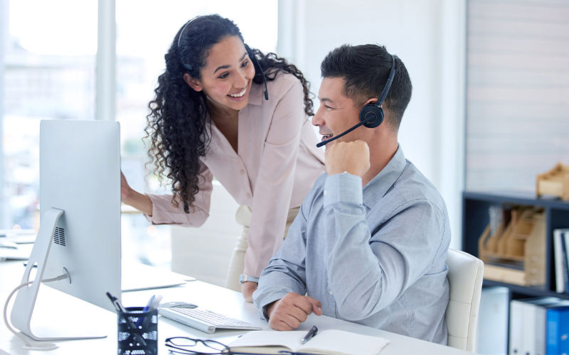 Two professionals collaborating in a modern office setting, discussing work at a desktop computer. One person is wearing a headset, and the workspace features bright natural lighting and minimalistic design.