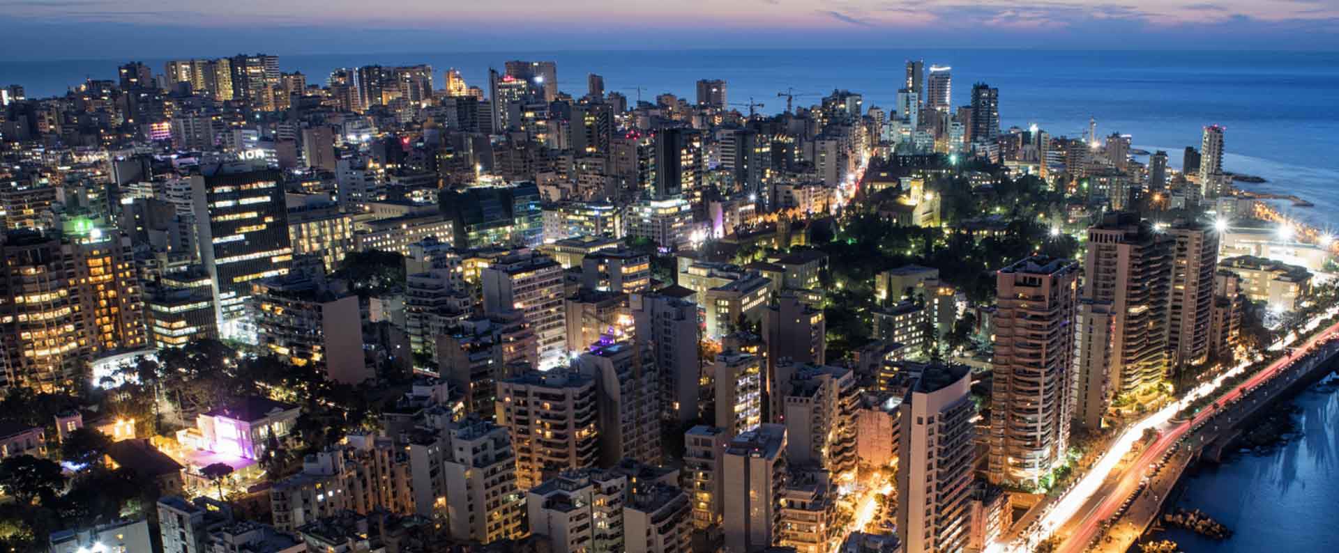 A dense cityscape with illuminated skyscrapers spreads along a coastline at dusk, reflecting light off the sea, creating a vibrant urban environment.