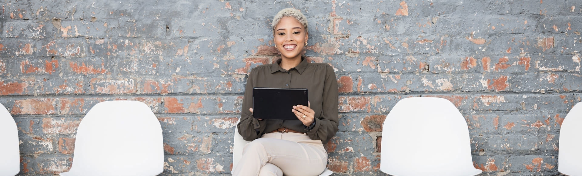Smiling Afro-descendant woman in business attire using a tablet during a job interview in an office with red brick walls.