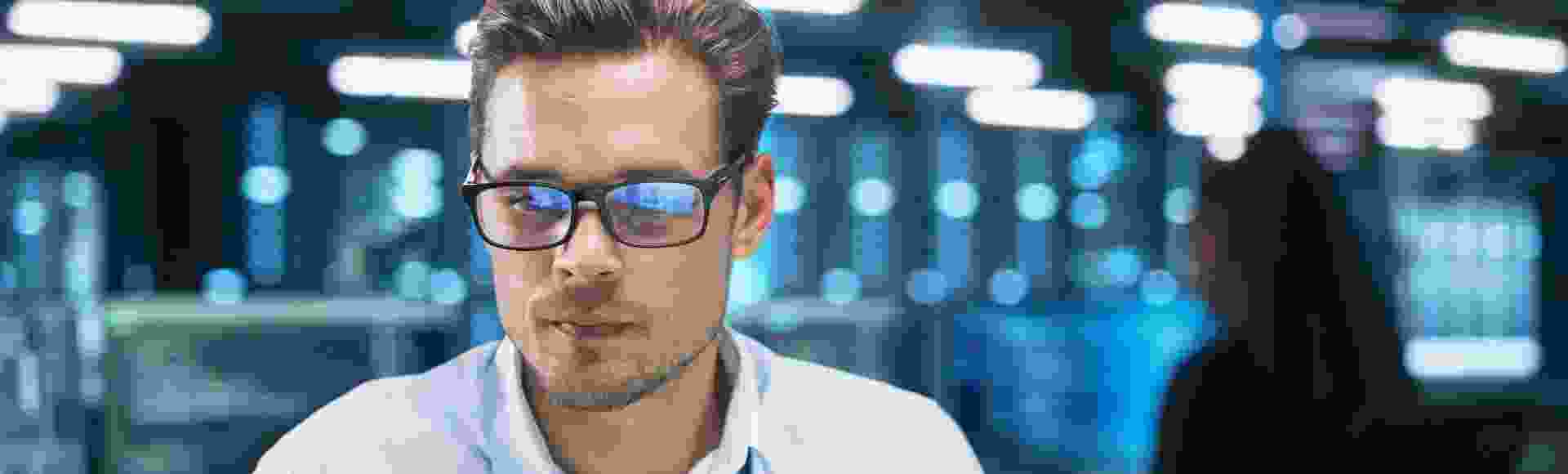 Man wearing glasses working inside a data center, surrounded by server racks and high-tech equipment.