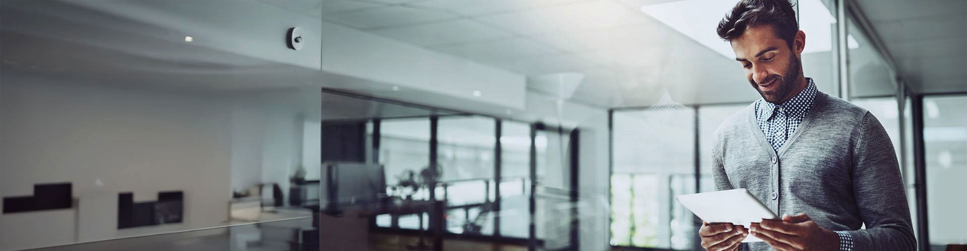 A person reads a tablet while standing in a modern, brightly-lit office with large windows and a reflective wall.