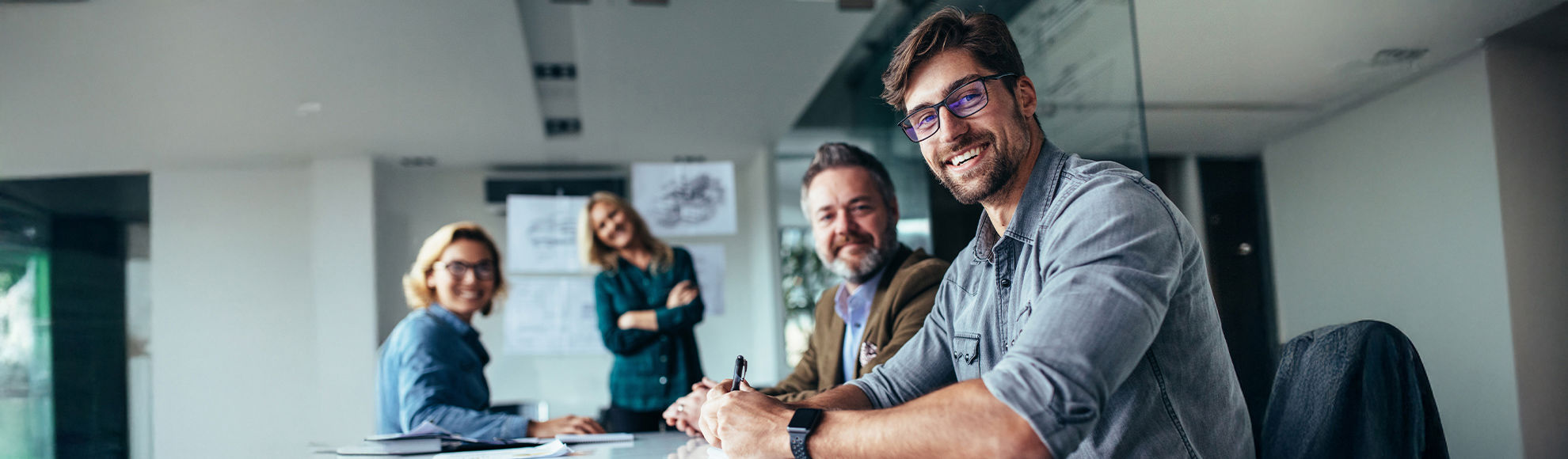 Four professionals in a modern office setting engaged in a collaborative meeting around a conference table, with glass walls and architectural drawings in the background.