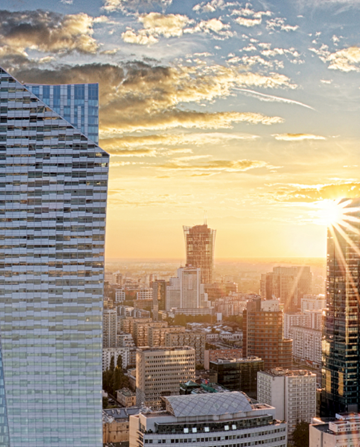 Panorama miasta o zachodzie słońca z wysokimi budynkami odbijającymi promienie słoneczne i mniejszymi konstrukcjami pod pochmurnym niebem.
