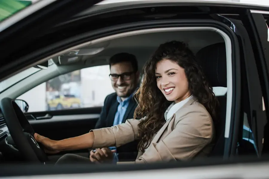 Two professionals smiling while sitting inside a car at a dealership showroom.