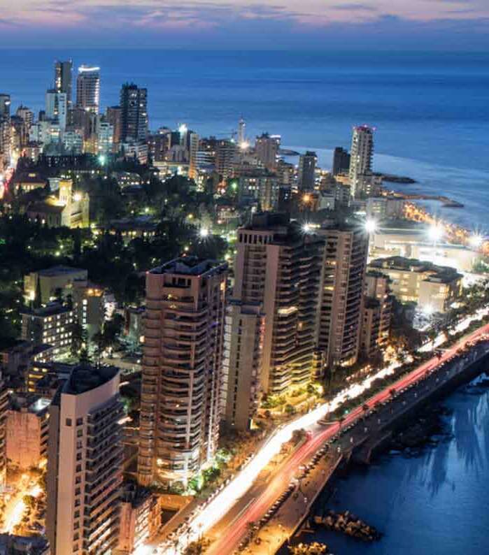 Buildings stand illuminated against a twilight sky, with a busy street below showing streaks of car lights. the ocean is visible on the right.