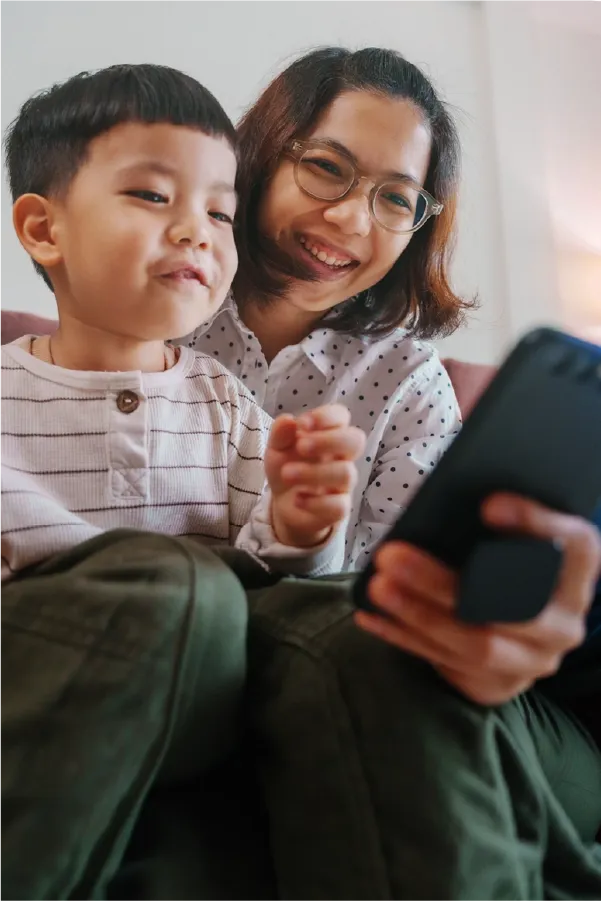 Mother and child smiling while using a smartphone together on a sofa.