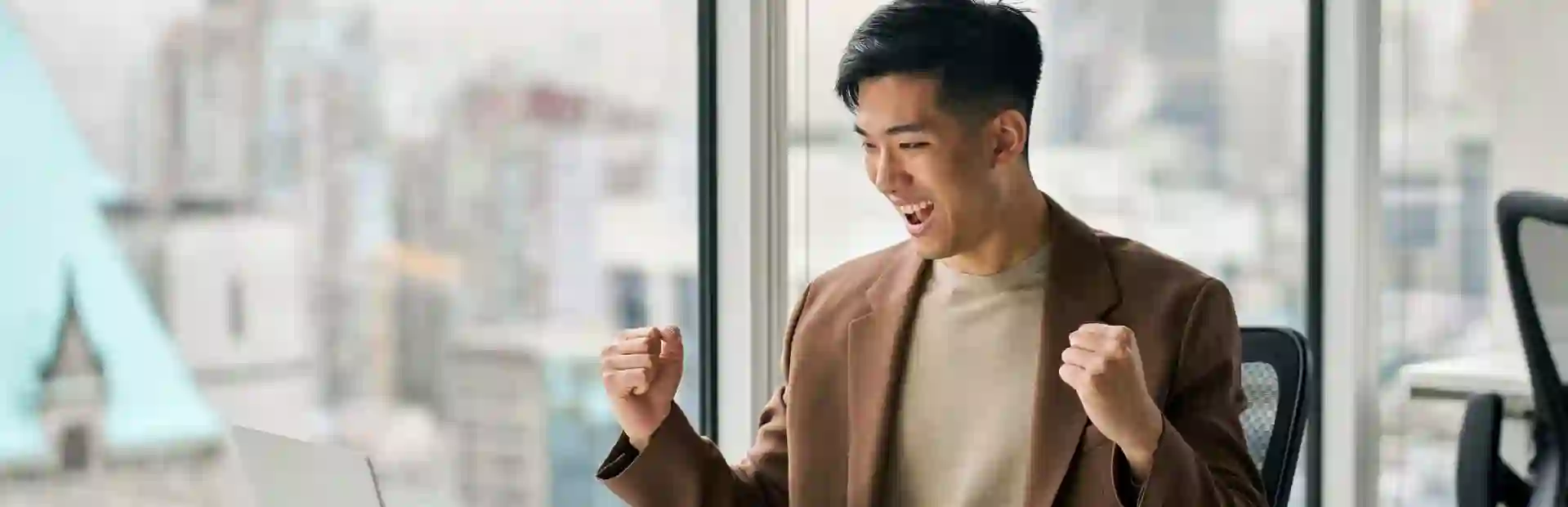 Asian man in a brown suit celebrates in front of his computer after receiving good news, inside a modern glass-walled office.