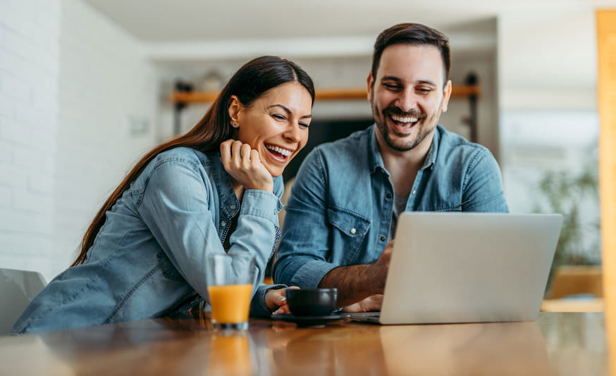 Relaxed couple at home viewing content on a laptop, with a cup of coffee and a glass of juice on the table.