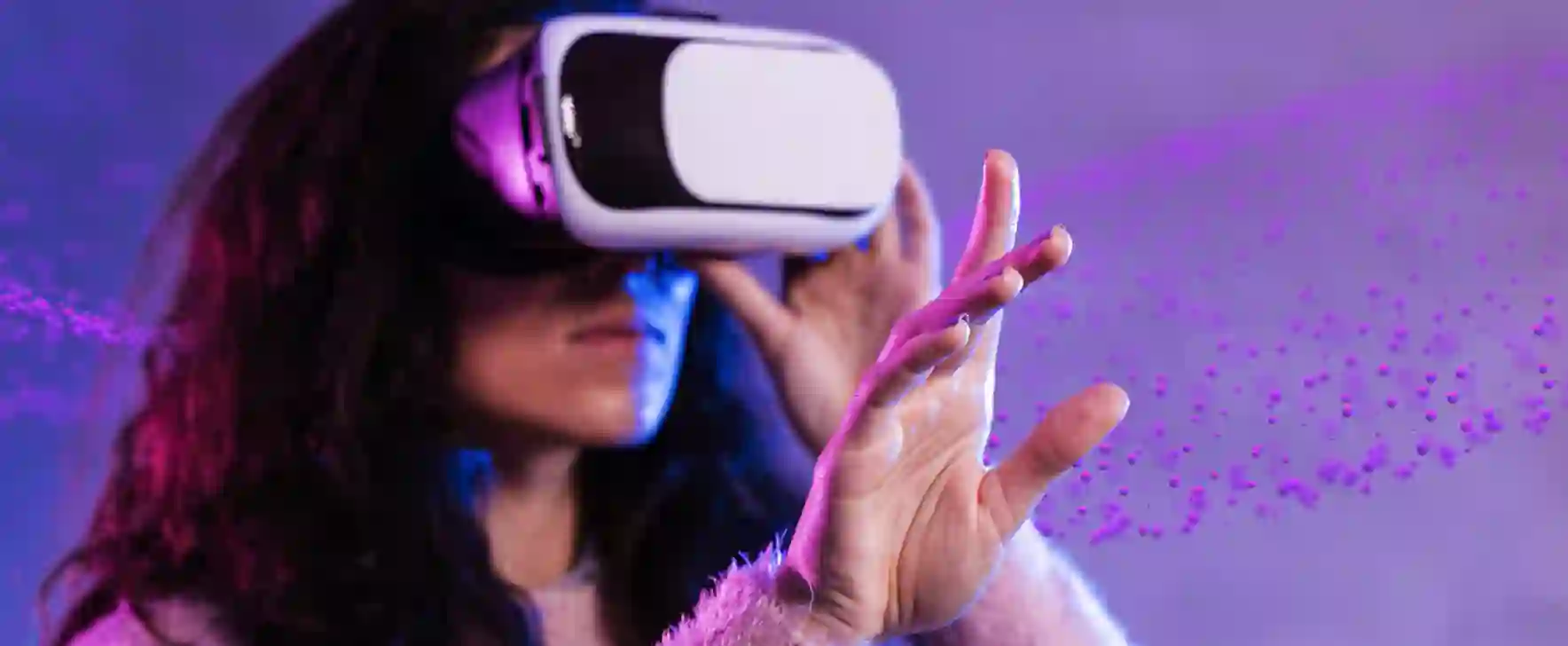 Close-up of a woman wearing virtual reality goggles, reaching forward with her hand, illuminated by purple and blue neon light effects.
