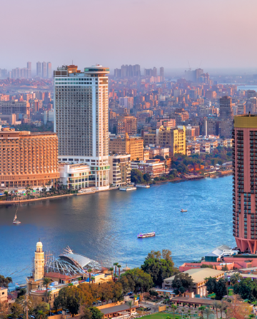 A tall modern skyscraper overlooks a river with a boat floating by, surrounded by densely packed buildings under a pinkish sky at dusk, creating an urban cityscape.