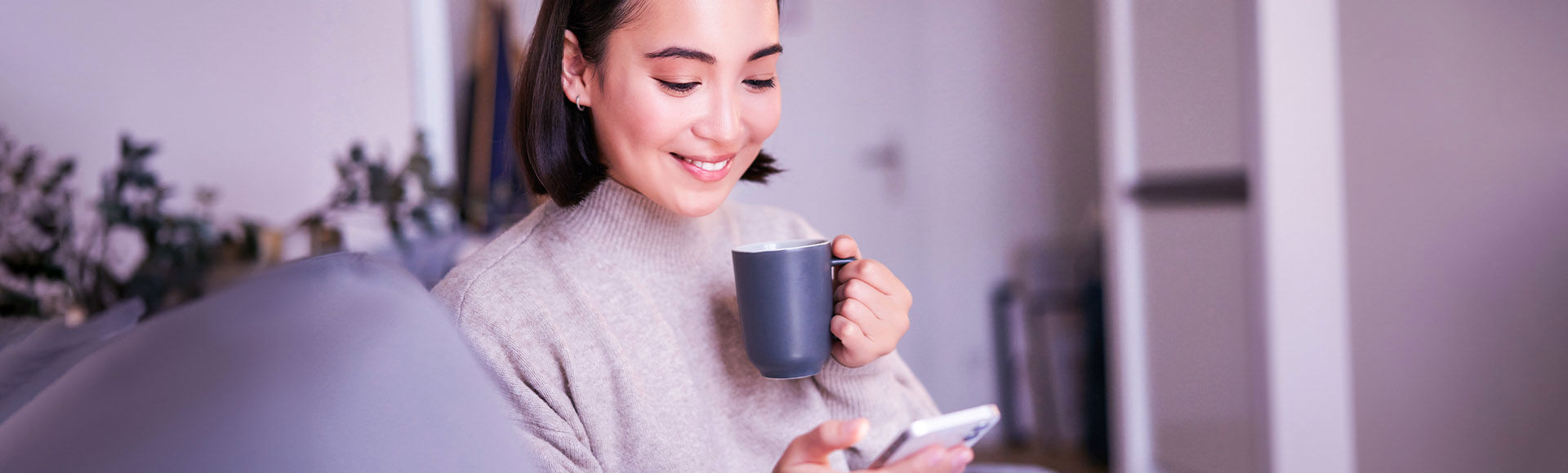 A smiling woman relaxes with coffee while using her smartphone at home.