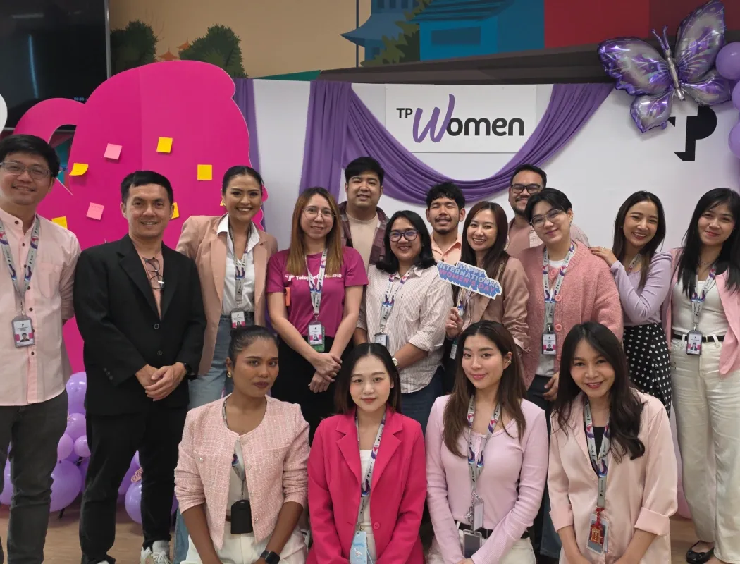 A group of TP employees poses cheerfully at a TP Women International Women's Day event, celebrating with a festive, decorated backdrop.