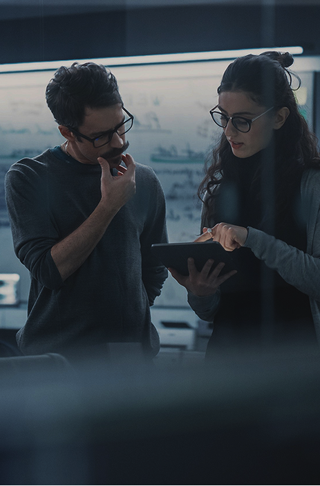 Two colleagues discussing while looking at a tablet in a dimly lit office.