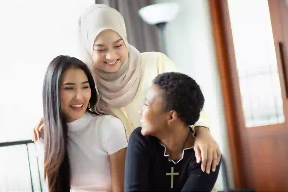 Three women smiling together indoors, one wearing a hijab, one with long straight hair, and one wearing a cross necklace, showing friendship and diversity.