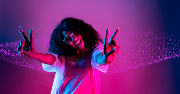 Smiling woman wearing sunglasses posing with two peace signs under vibrant pink and purple neon lights