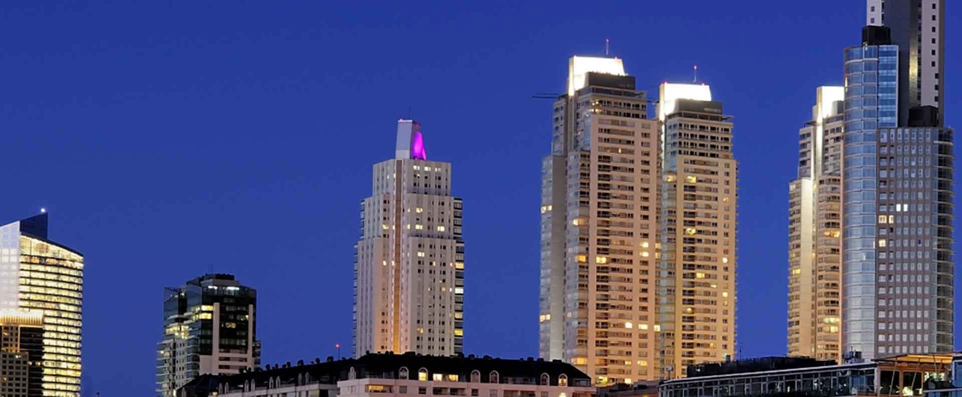 Altos rascacielos iluminados de noche contra un cielo azul profundo, con luces brillantes y edificios más pequeños abajo.