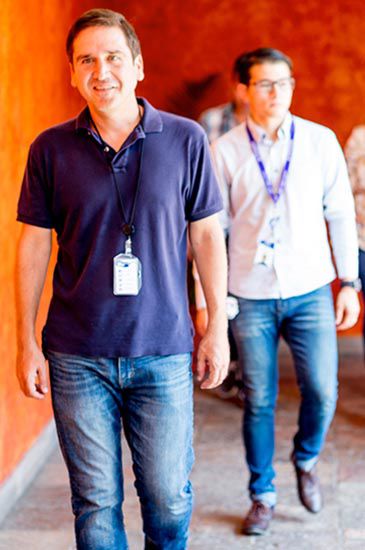 Una persona con polo azul y jeans camina hacia adelante, con una placa de identificación. Otra persona lo sigue con camisa blanca, ambos en un pasillo bien iluminado con paredes naranjas.