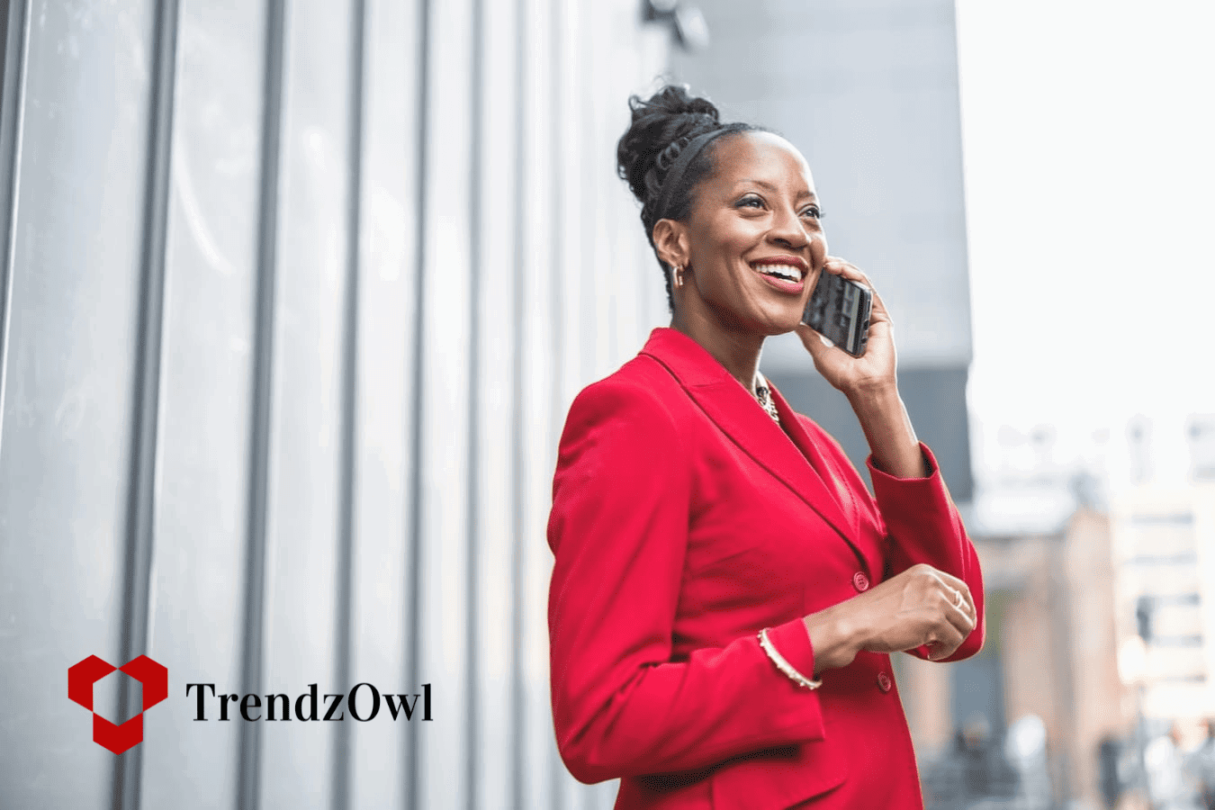 A woman in a bright red suit talks on a smartphone, smiling outdoors near a modern building. text: "trendzowl" with a red geometric heart logo. A woman in a bright red suit talks on a smartphone, smiling outdoors near a modern building. text: "trendzowl" with a red geometric heart logo.