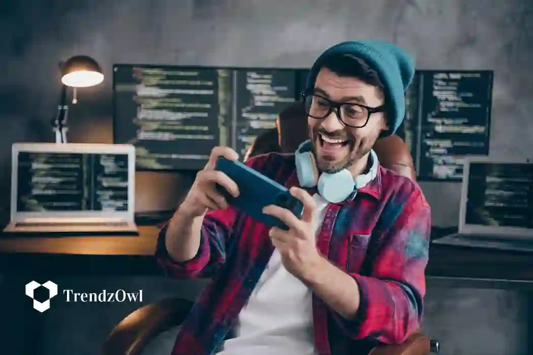 Young smiling gamer playing a game on his phone, wearing a headset around his neck in a room with multiple monitors.
