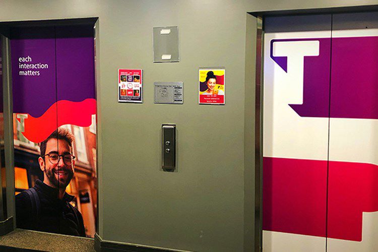 Two elevator doors are in a hallway. the left door features a smiling man and text: "each interaction matters." bright posters are on the wall between the elevators.