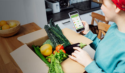 Person reviewing an online grocery app on a smartphone next to a box of fresh vegetables.