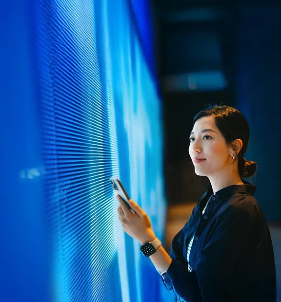 Asian woman holding a smartphone in front of a modern digital display.