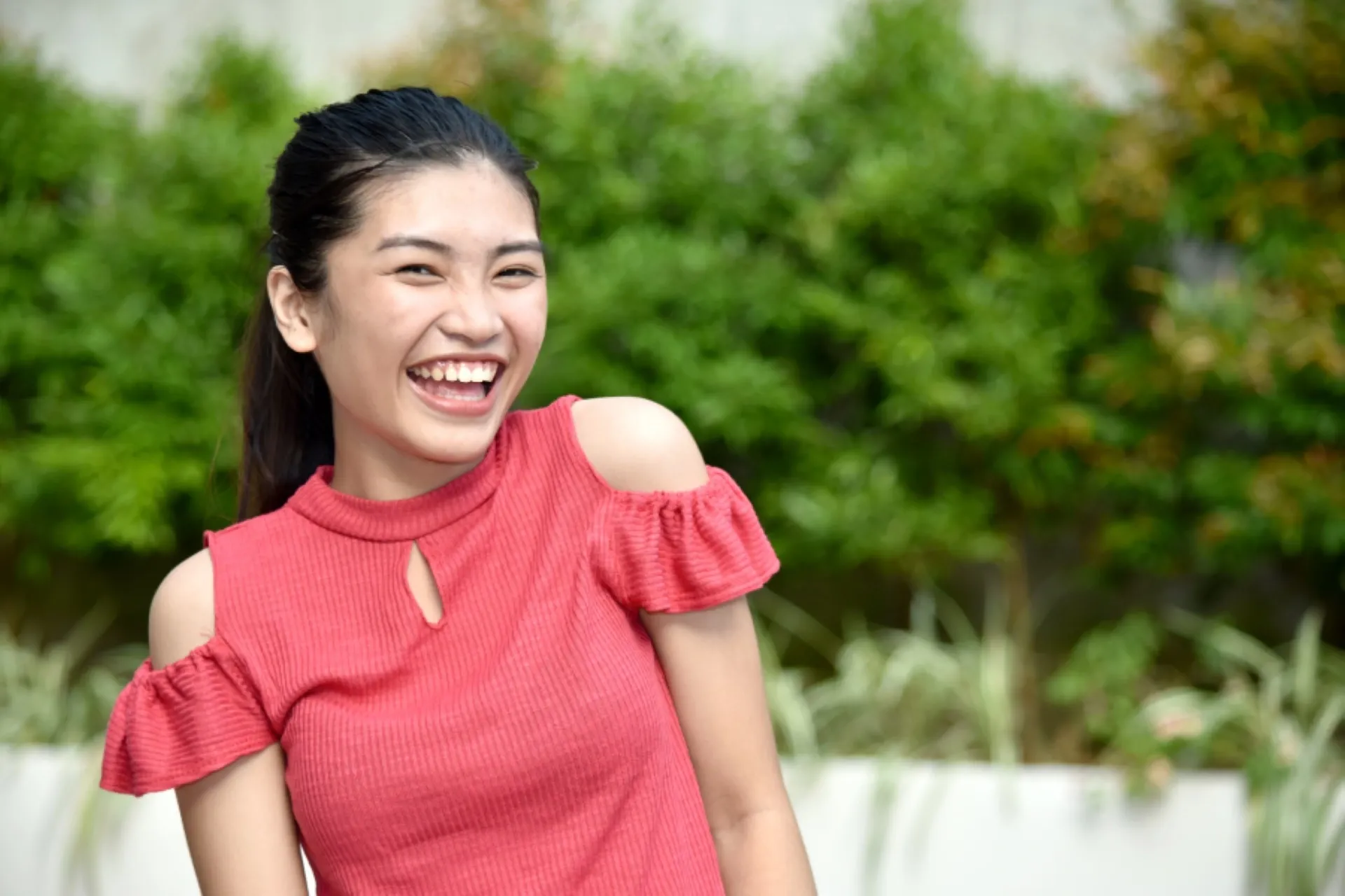 Young woman smiling outdoors in a red top with greenery in the background.