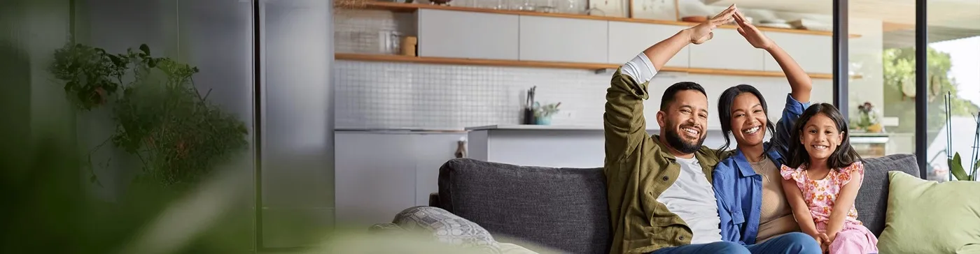 Smiling family sitting on a sofa at home, with parents forming a house gesture, representing TP’s Insurance page.