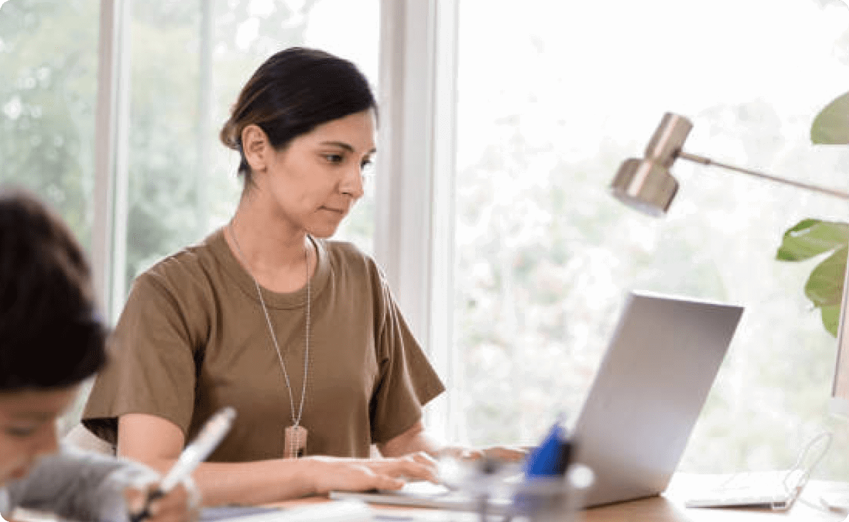 Woman typing on a laptop at a wooden desk, with a lamp nearby and a window showing greenery outside; a child is writing in the foreground.
