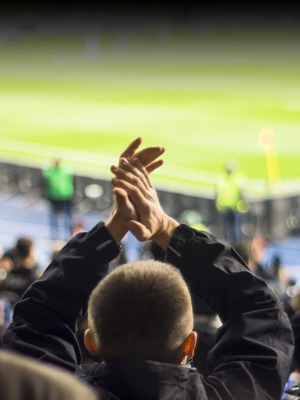 A person in a black jacket, clapping with hands raised, watches a sports event in a brightly-lit stadium