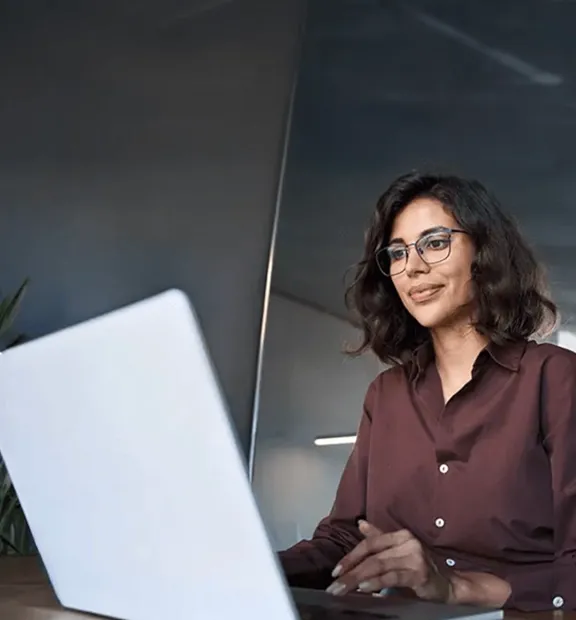 Businesswoman wearing glasses and a brown blouse working on her laptop in a modern office with glass walls.