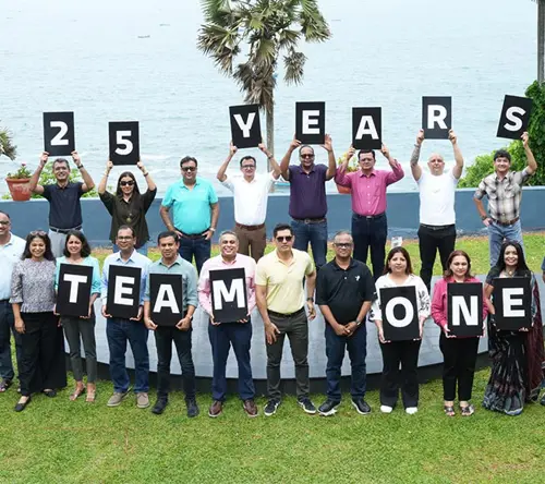 Team members celebrating TP's 25th anniversary by holding letter boards spelling '25 Years One Team One TP' at an outdoor waterfront event.