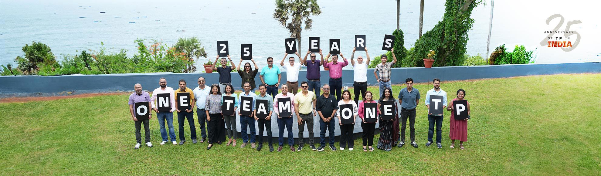 Team members celebrating TP's 25th anniversary by holding letter boards spelling '25 Years One Team One TP' at an outdoor waterfront event.
