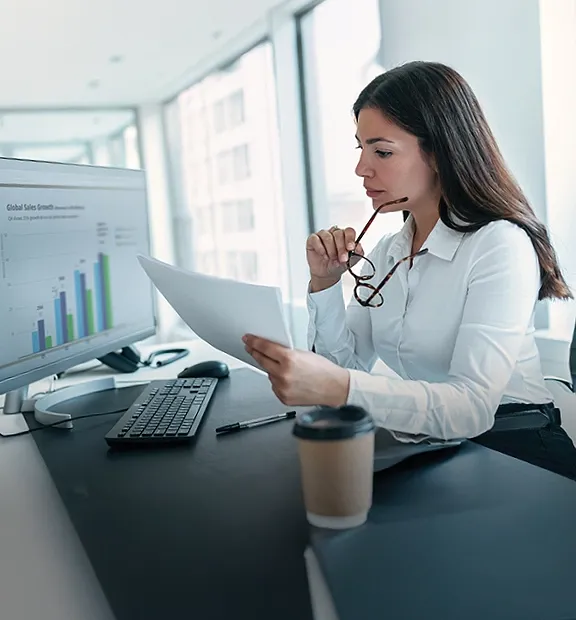 Businesswoman reviewing a report while analyzing sales data on a computer screen.