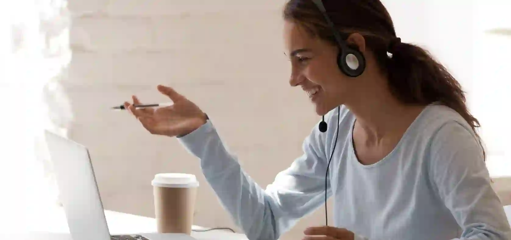 Smiling woman wearing a headset, talking and gesturing during a video call on a laptop with a coffee cup on the desk